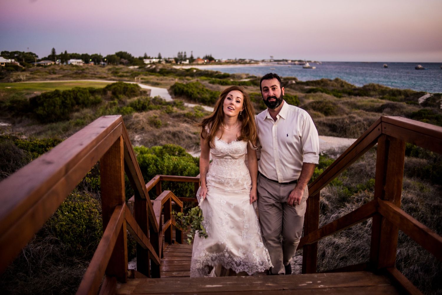 A couple in wedding attire poses on wooden stairs overlooking a coastal landscape at sunset.