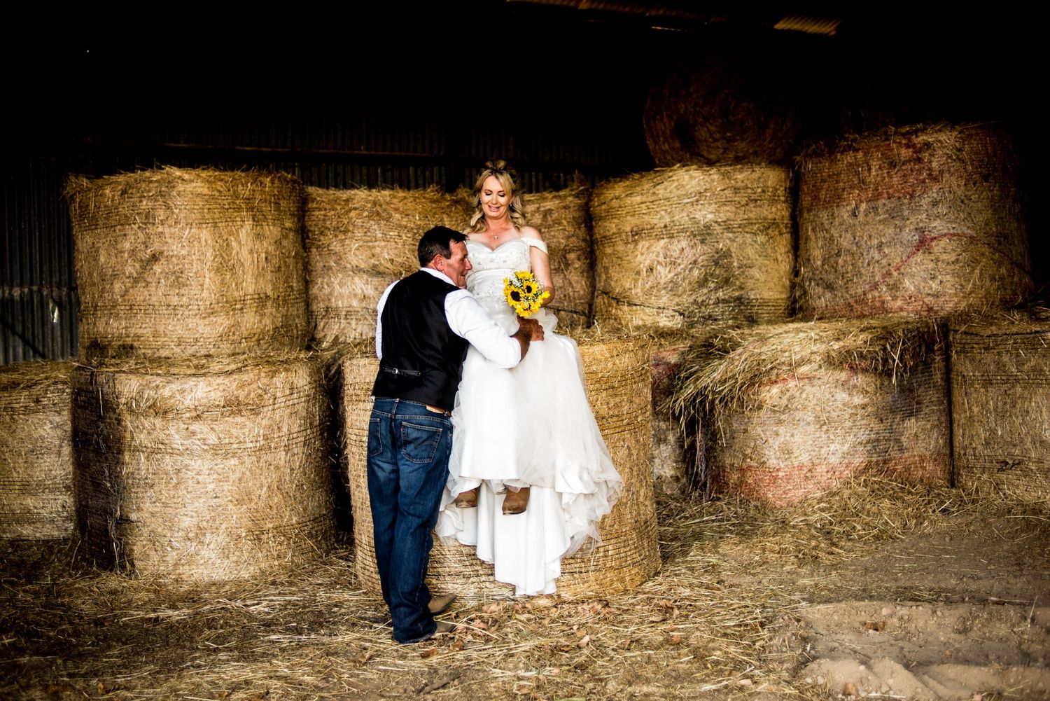 A couple poses against stacked hay bales in a rustic barn setting.