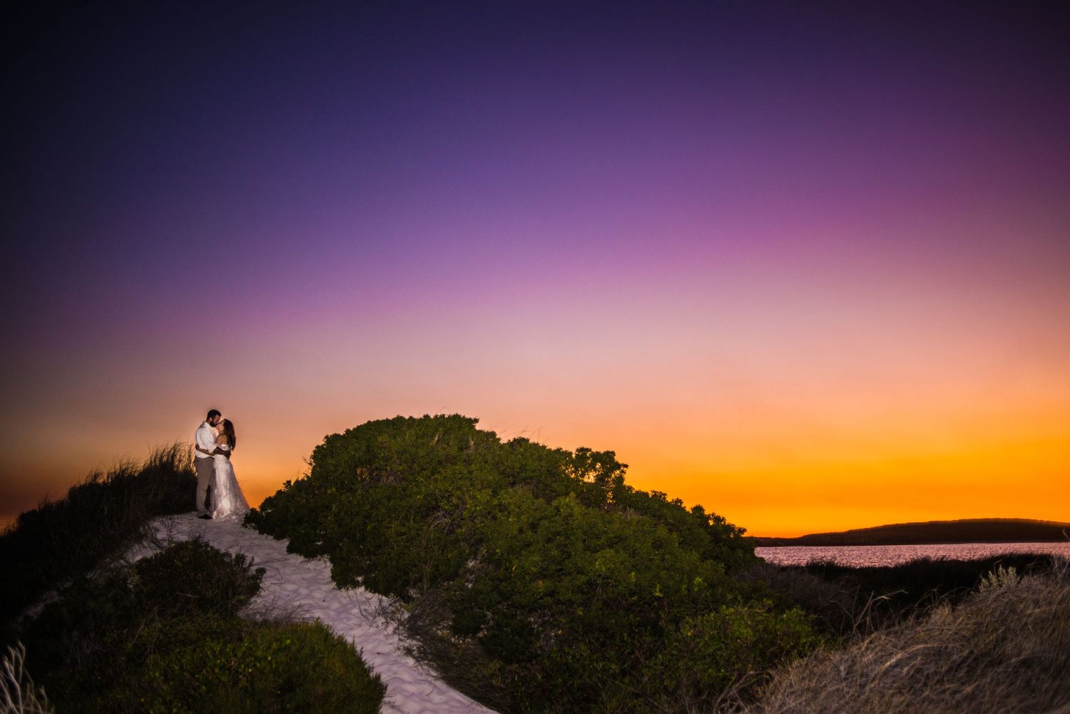 A couple stands silhouetted against a vibrant purple and orange sunset sky on a hilltop path.