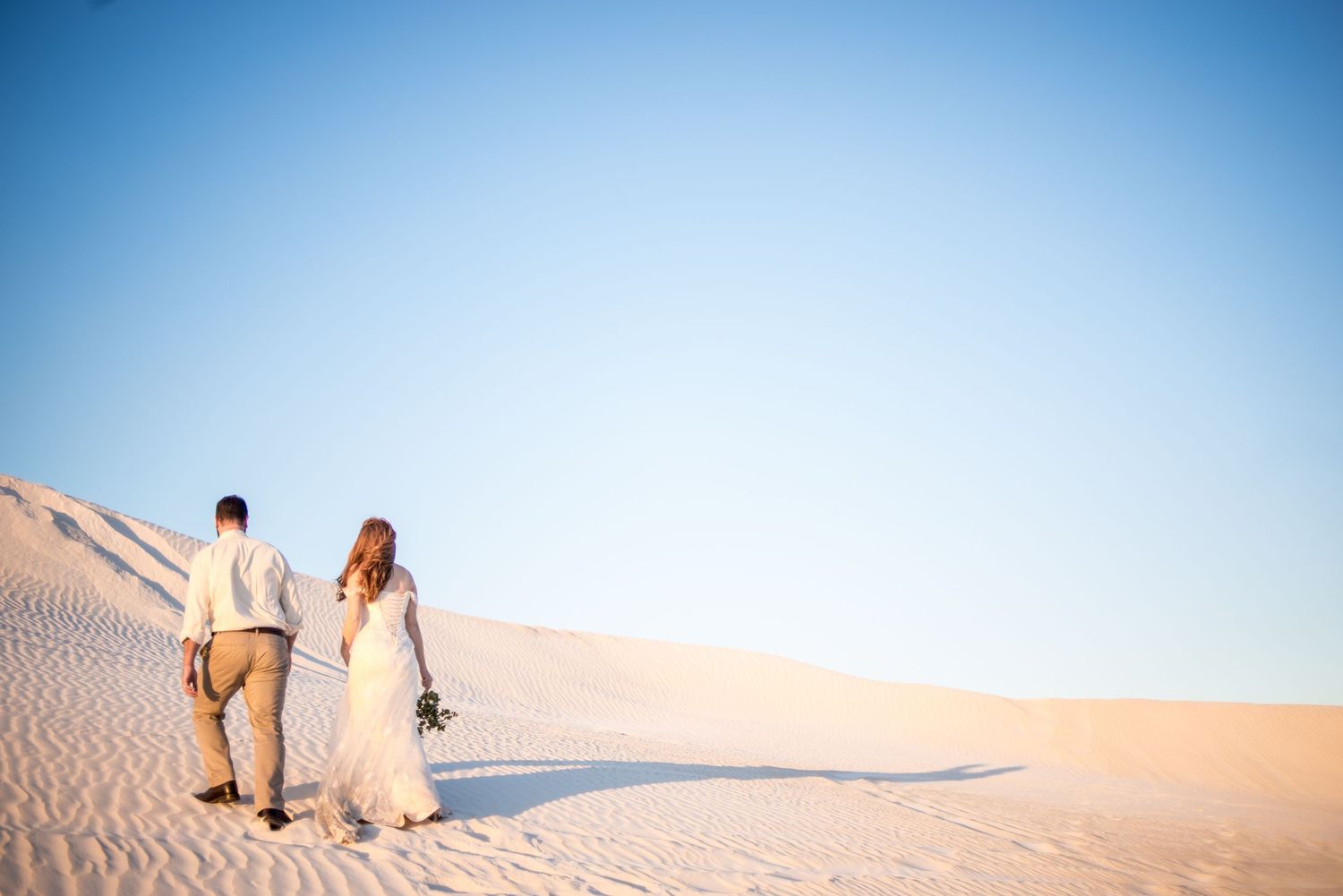 A couple walks hand in hand across white sand dunes under clear blue skies.