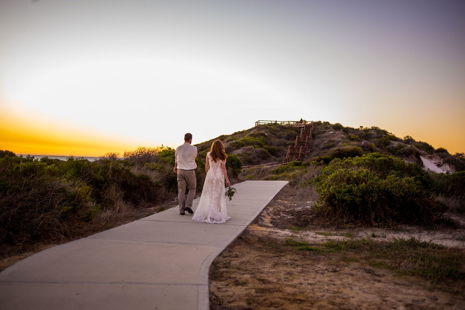 A couple in wedding attire walks along a coastal path during golden hour.