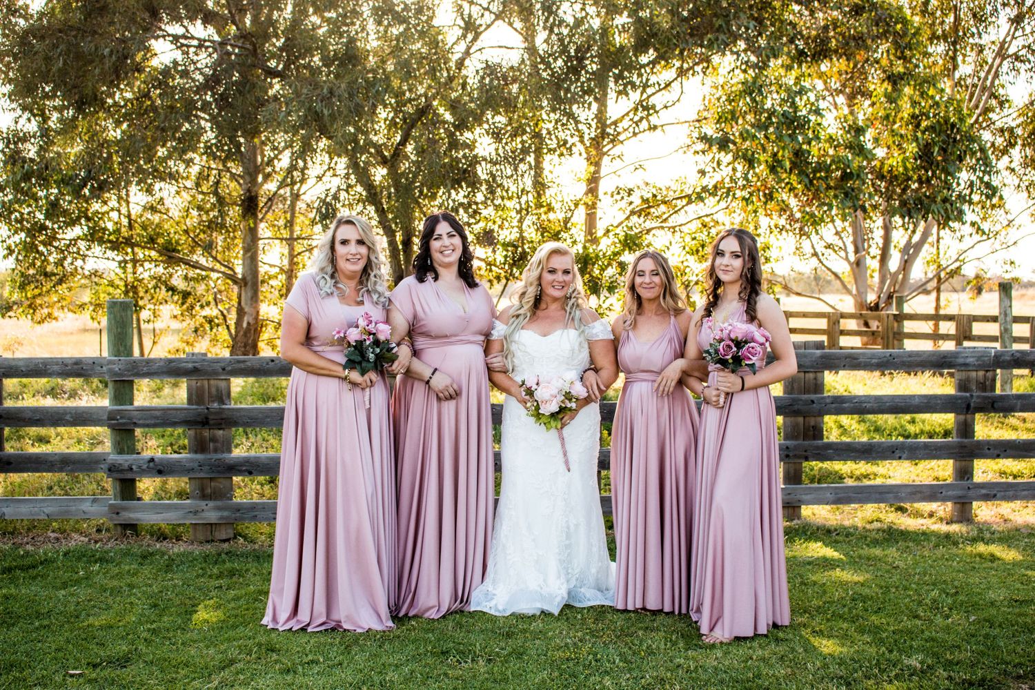 A bridal party poses in a garden setting wearing matching pink dresses against a wooden fence.