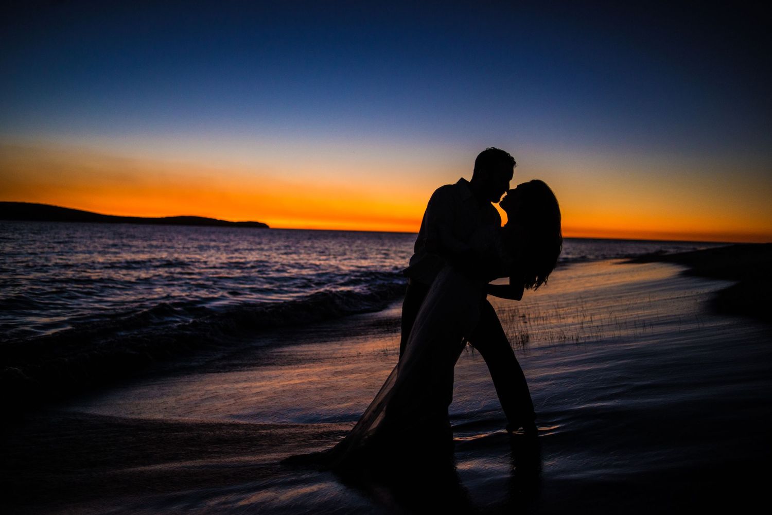 A romantic silhouette of a couple dips on the beach against a dramatic sunset backdrop.