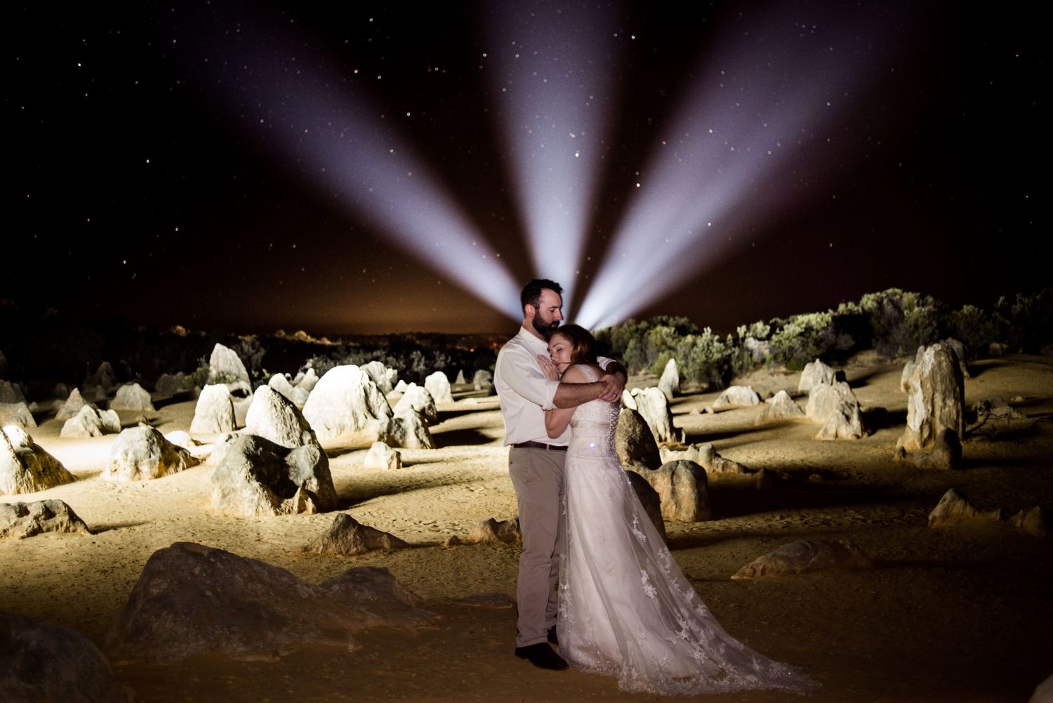 A couple poses in wedding attire against dramatic spotlights in a unique rock formation setting.