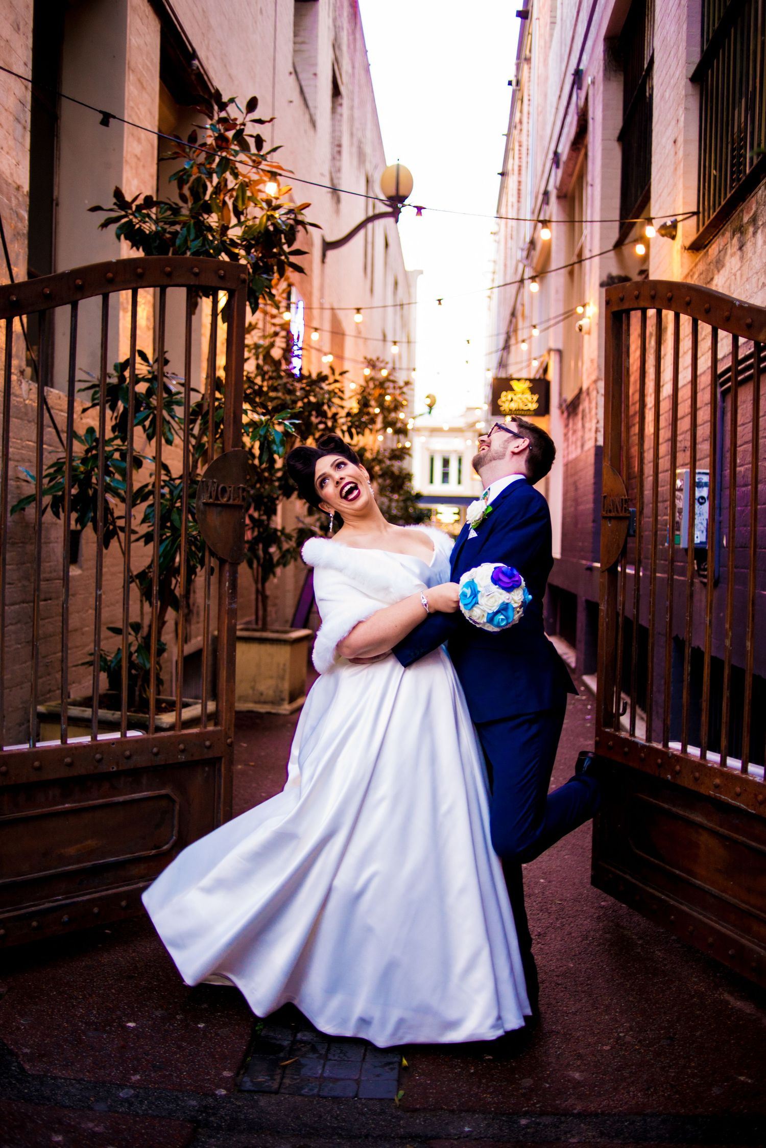 Bride and groom dance playfully in an urban alleyway with string lights overhead.