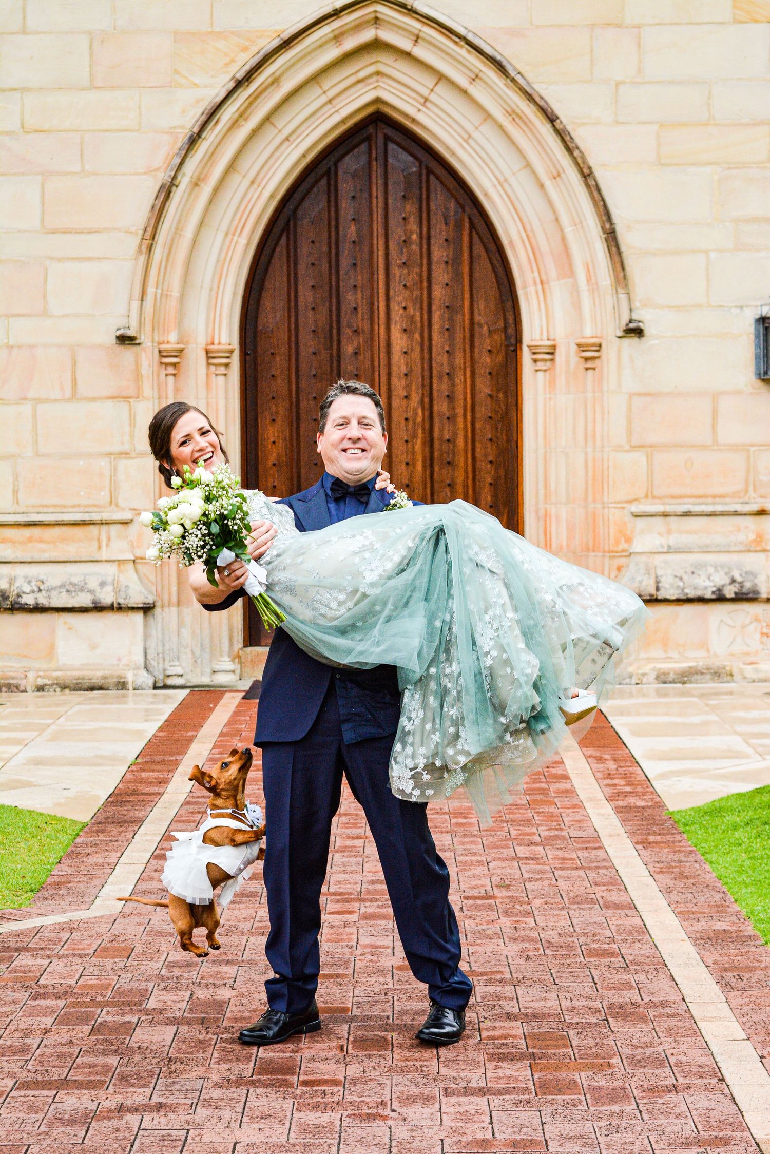 A couple poses with their dog in front of a gothic church doorway in a traditional wedding photo.