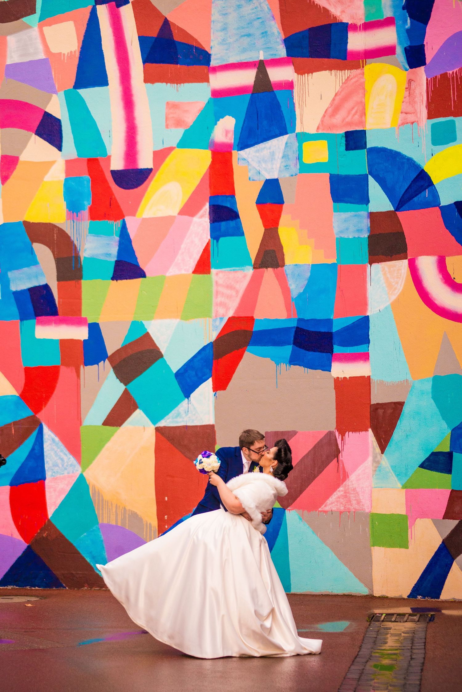 A couple dances in front of a vibrant geometric wall mural in wedding attire.