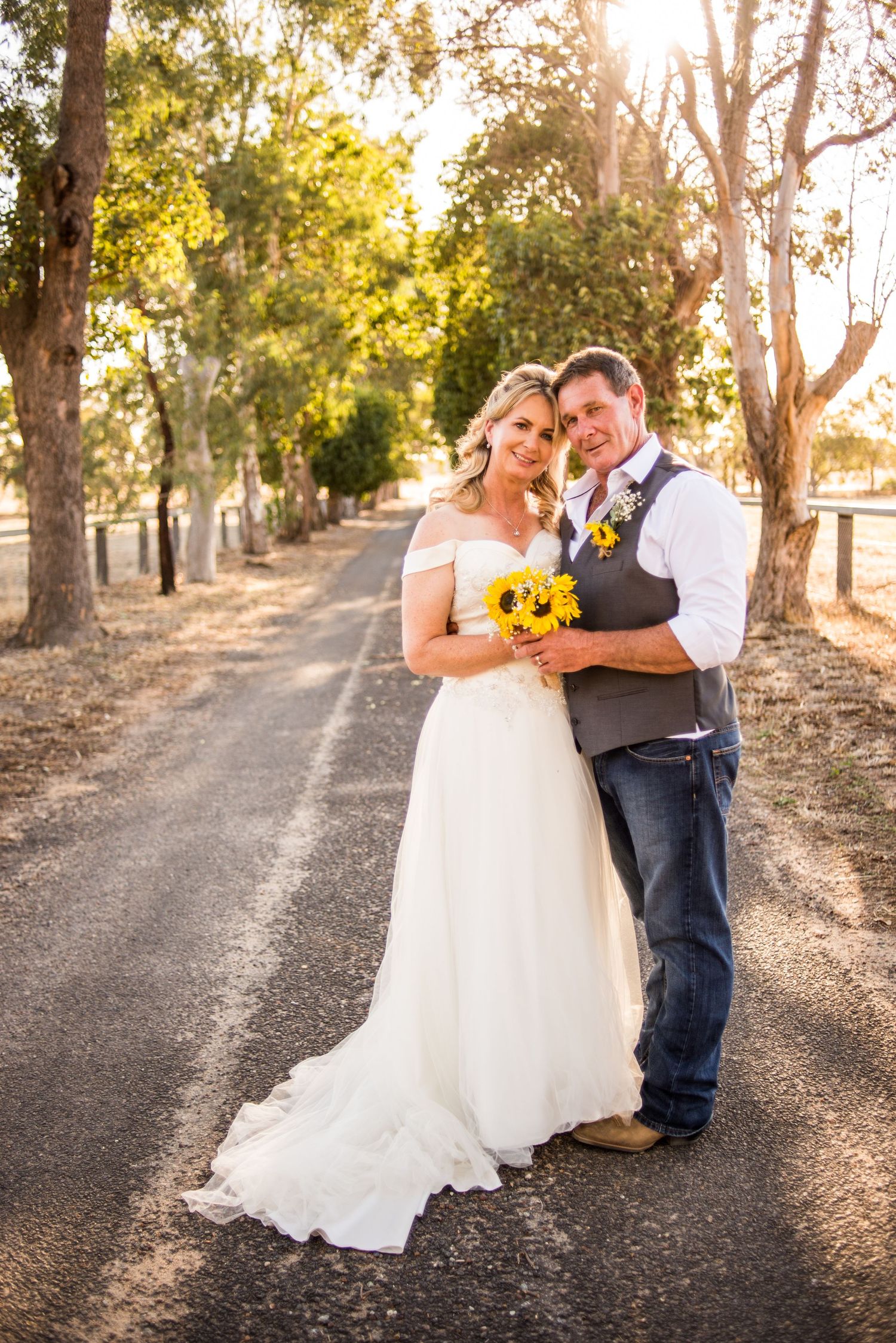A couple poses on a tree-lined country road holding sunflowers in rustic wedding attire.