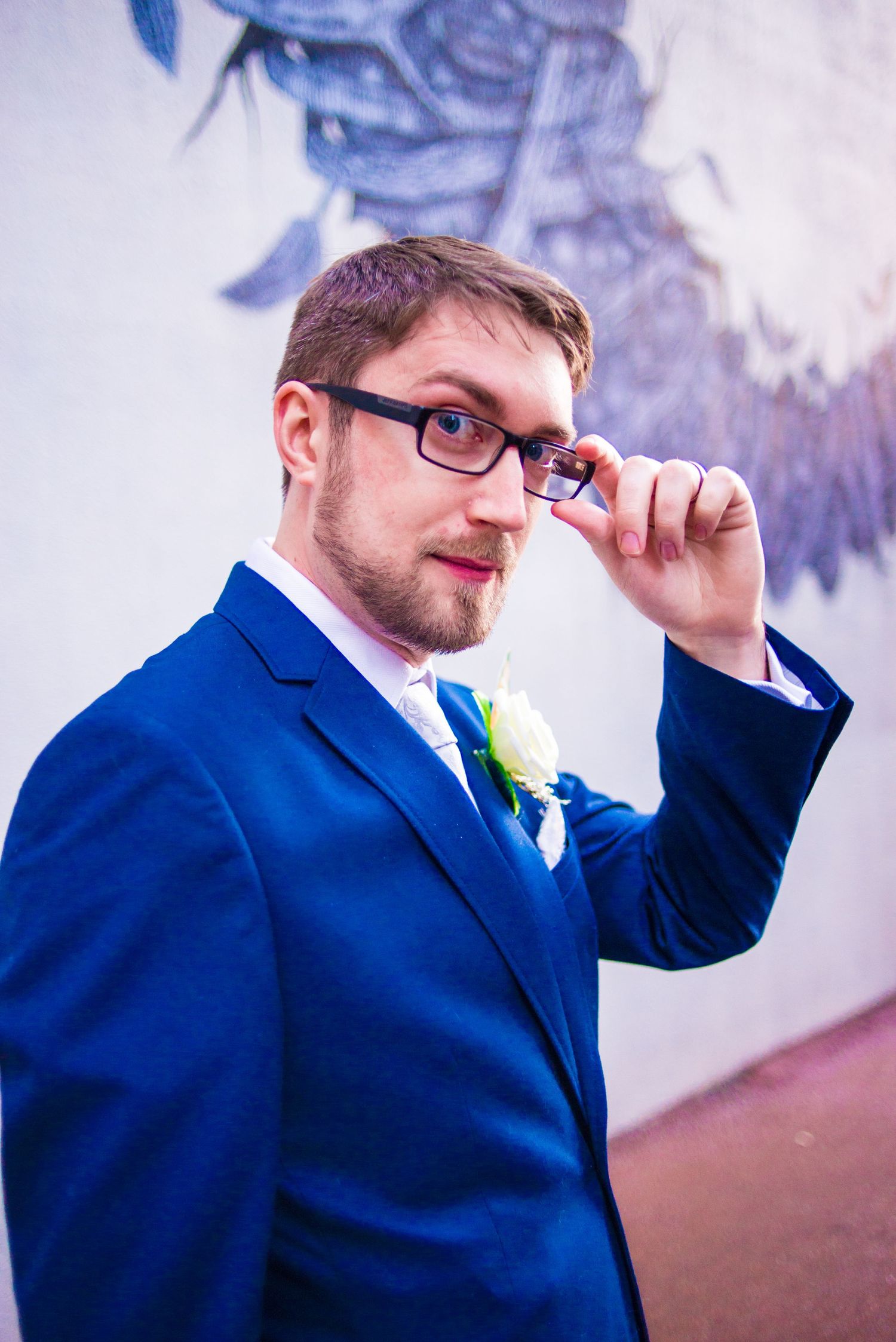 A groom in a navy blue suit adjusts his glasses while wearing a boutonniere.