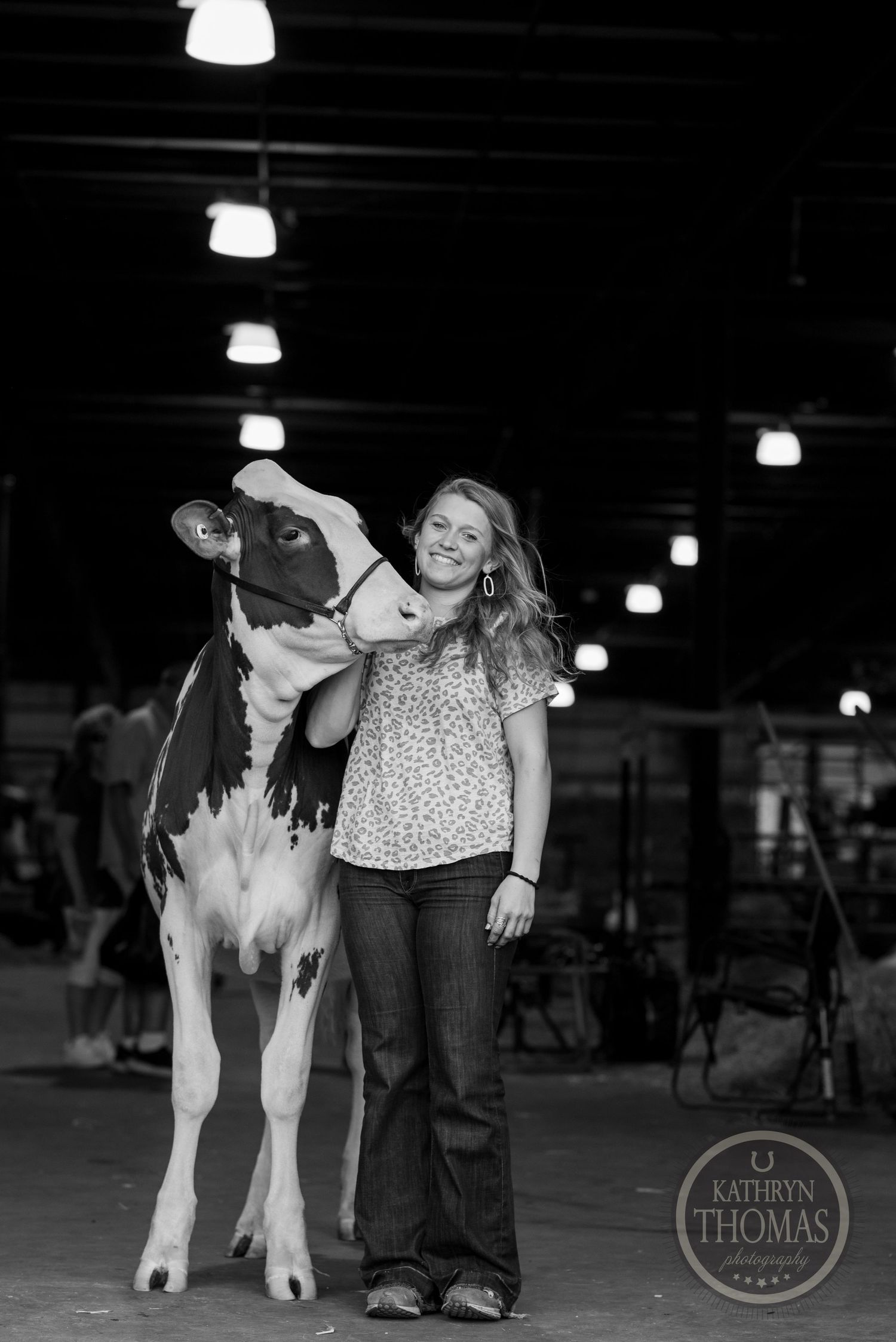 A black and white indoor photo shows a dairy cow and handler under barn lighting.