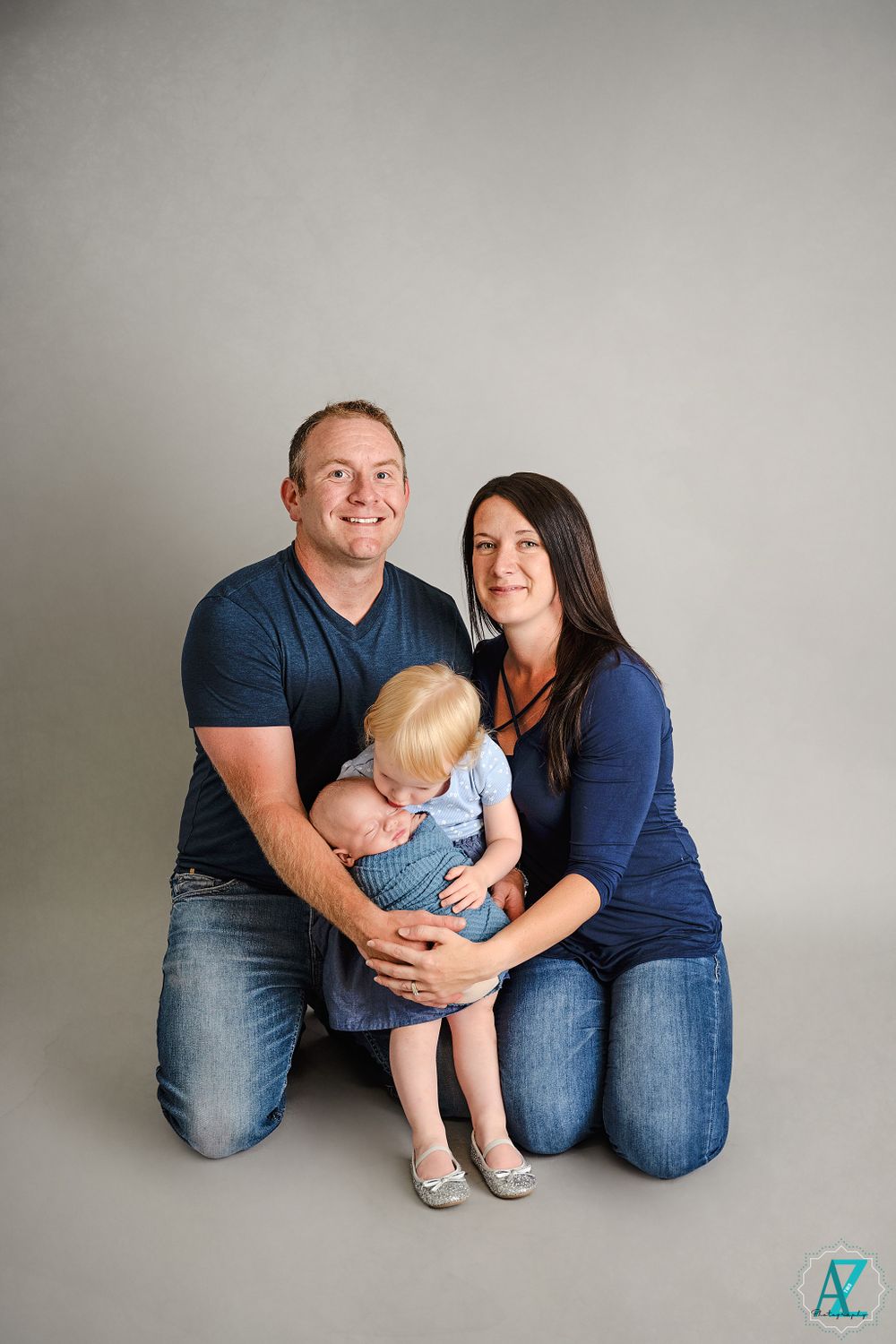 Family portrait with parents and toddler in coordinating blue outfits against grey backdrop.