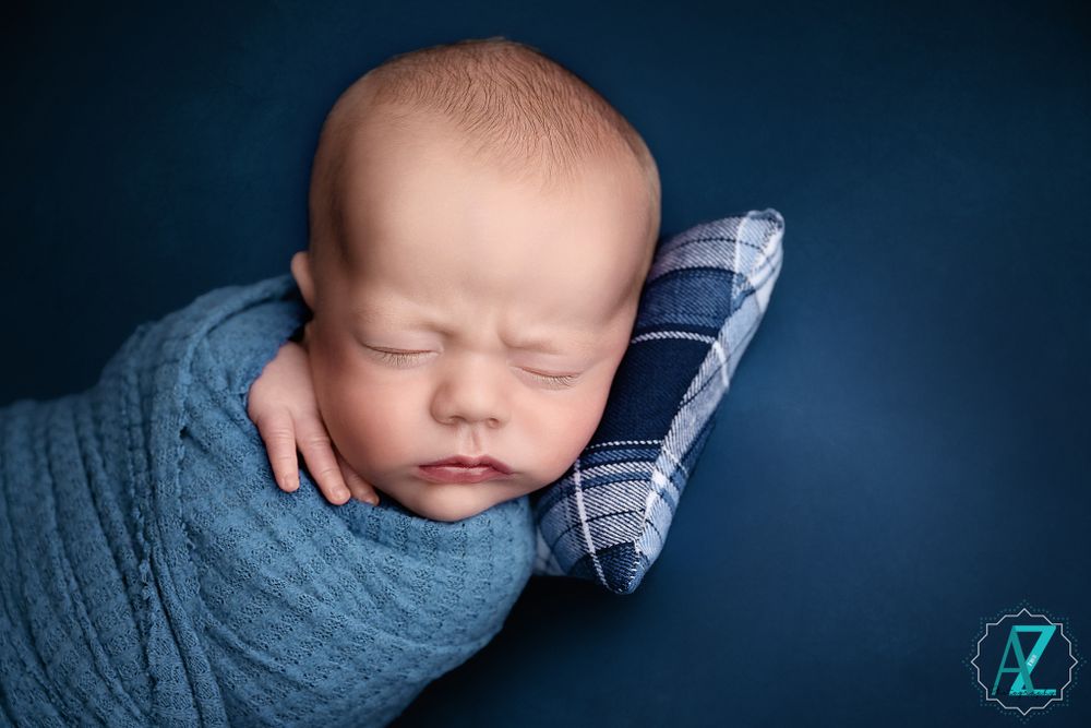 Newborn baby wrapped in teal knit blanket sleeping peacefully against navy blue background.