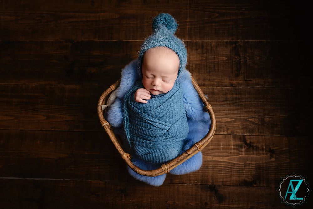 Peaceful sleeping baby in blue knit wrap and hat resting in woven basket against wooden backdrop.