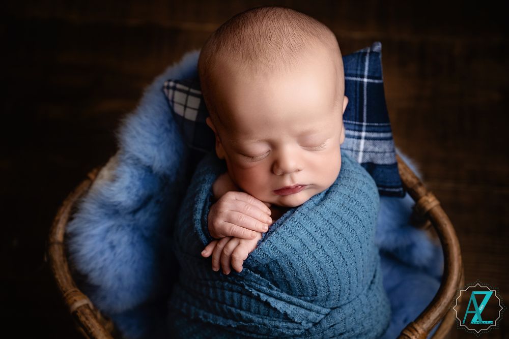 Sleeping newborn in blue wrap nestled in basket with plaid accent and fuzzy blue padding.