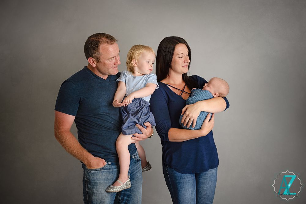 Family portrait showing parents with toddler and newborn against grey background.