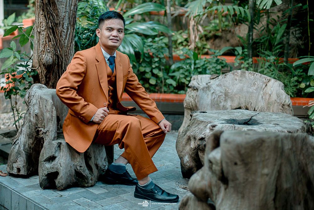 Groom in burnt orange suit sits on rustic wooden bench in tropical garden setting.