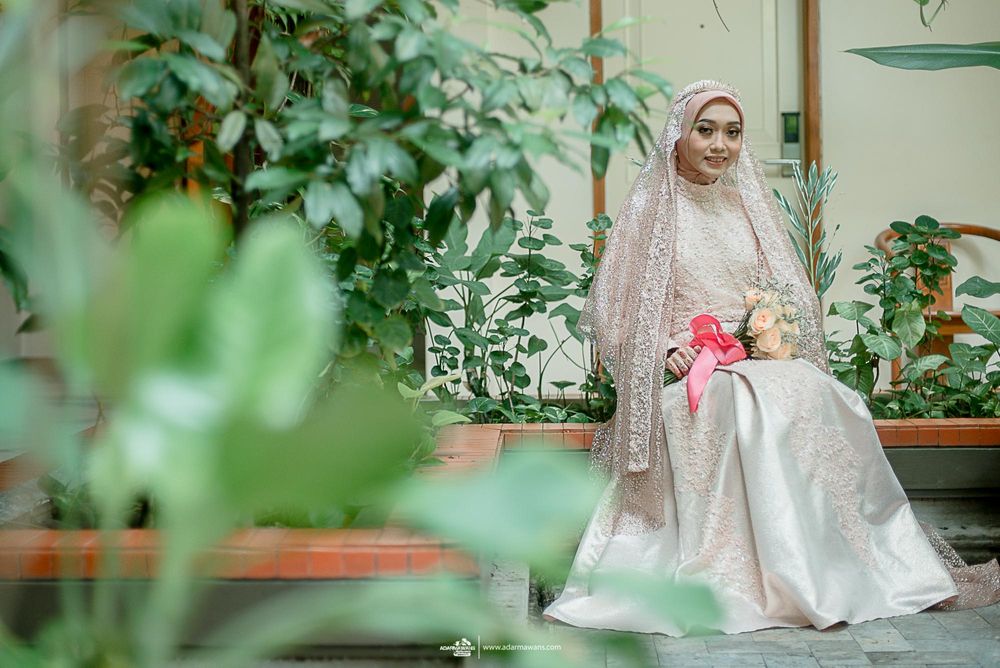 Bride in elegant white gown sits in lush garden setting with pink floral accents.