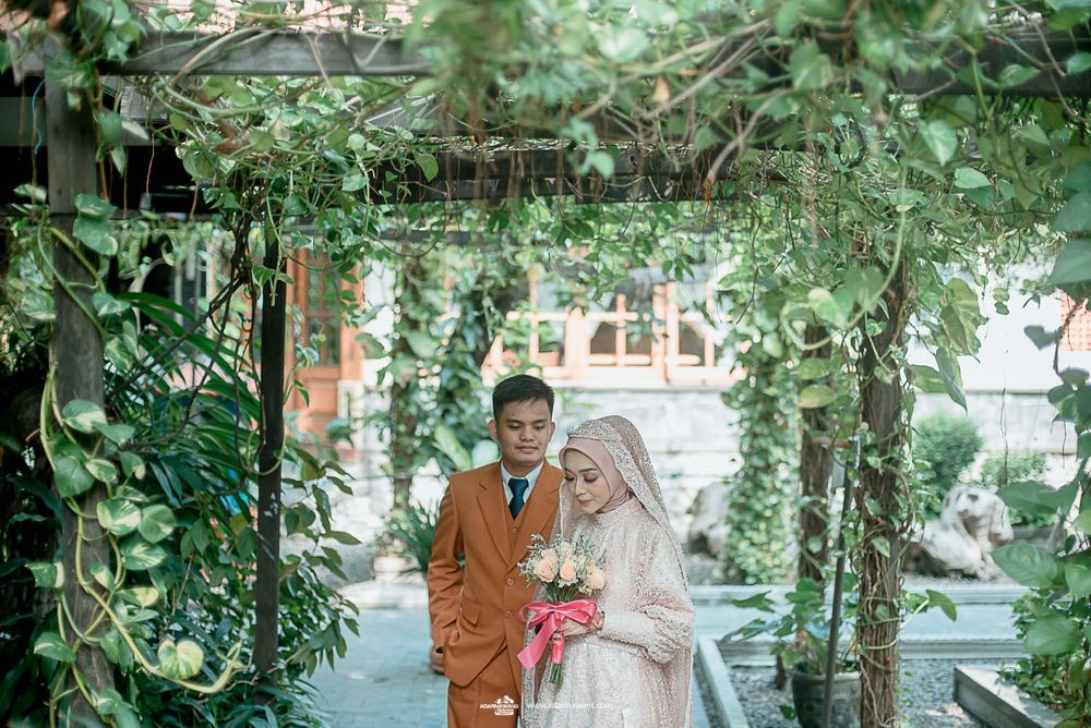 A couple stands beneath a lush green archway of vines and leaves in an outdoor garden setting.