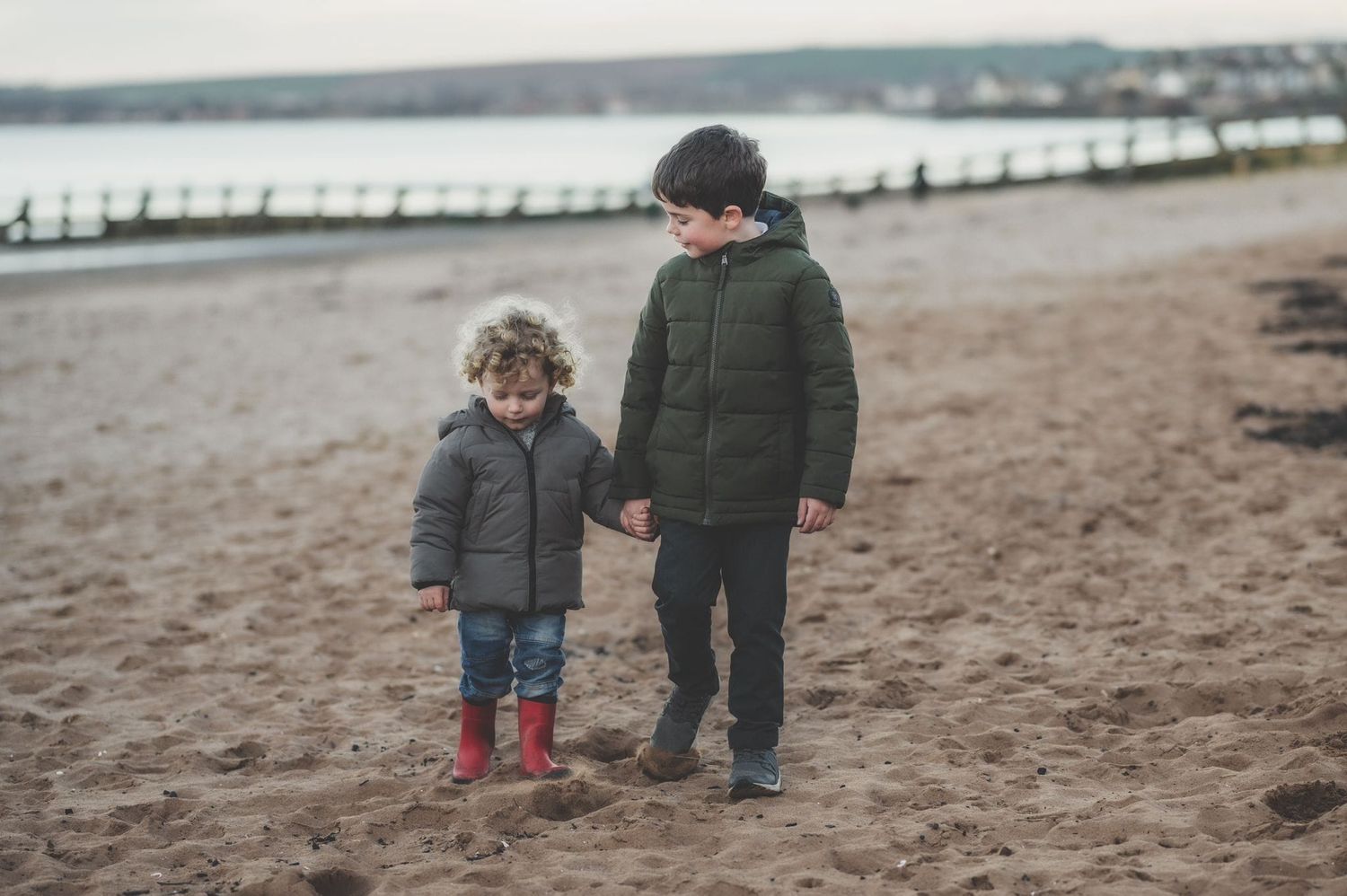 Two people walking together on a beach in winter coats near a pier.
