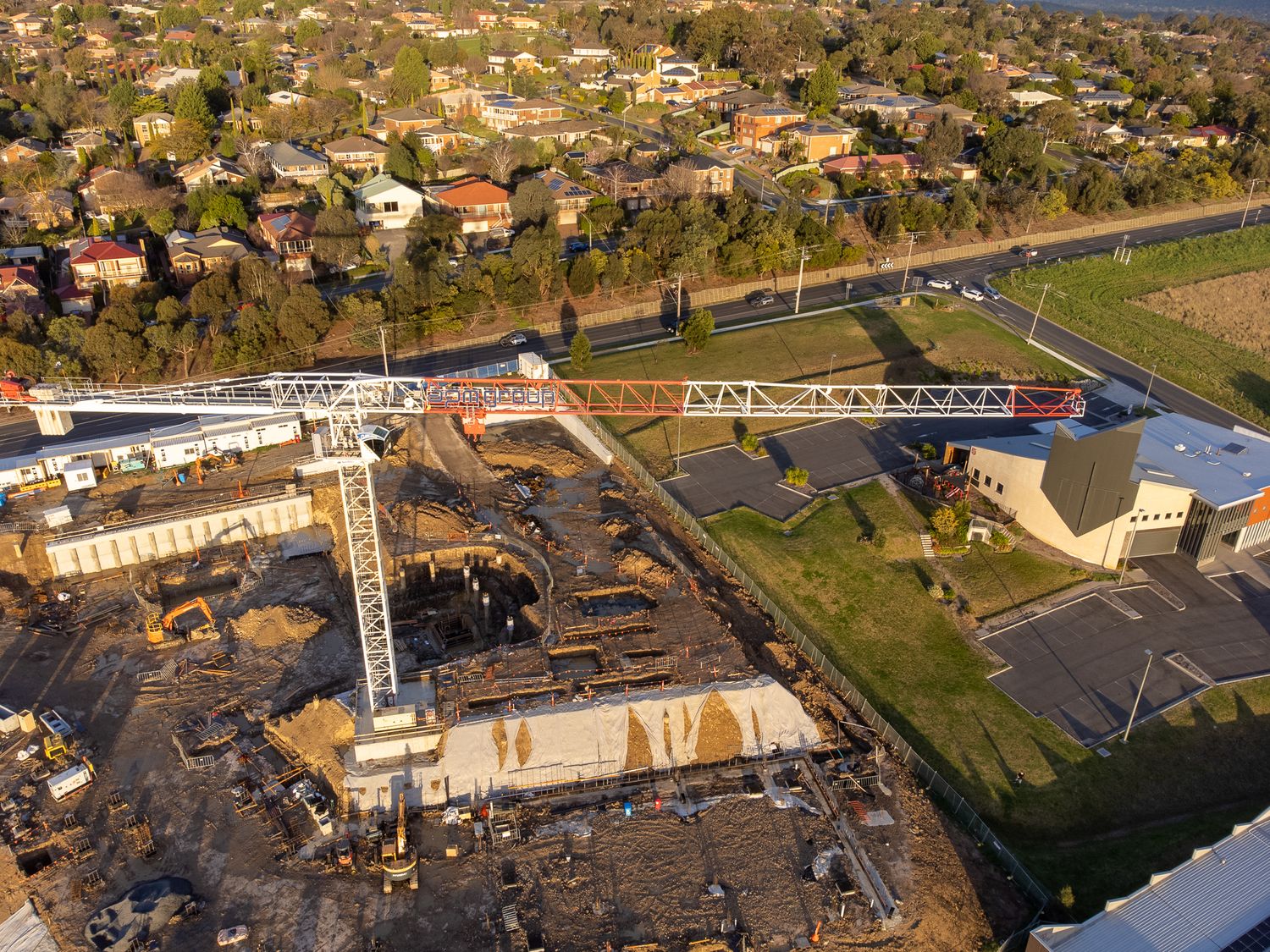 Aerial view of large suburban construction site with cranes near residential neighborhood and road.
