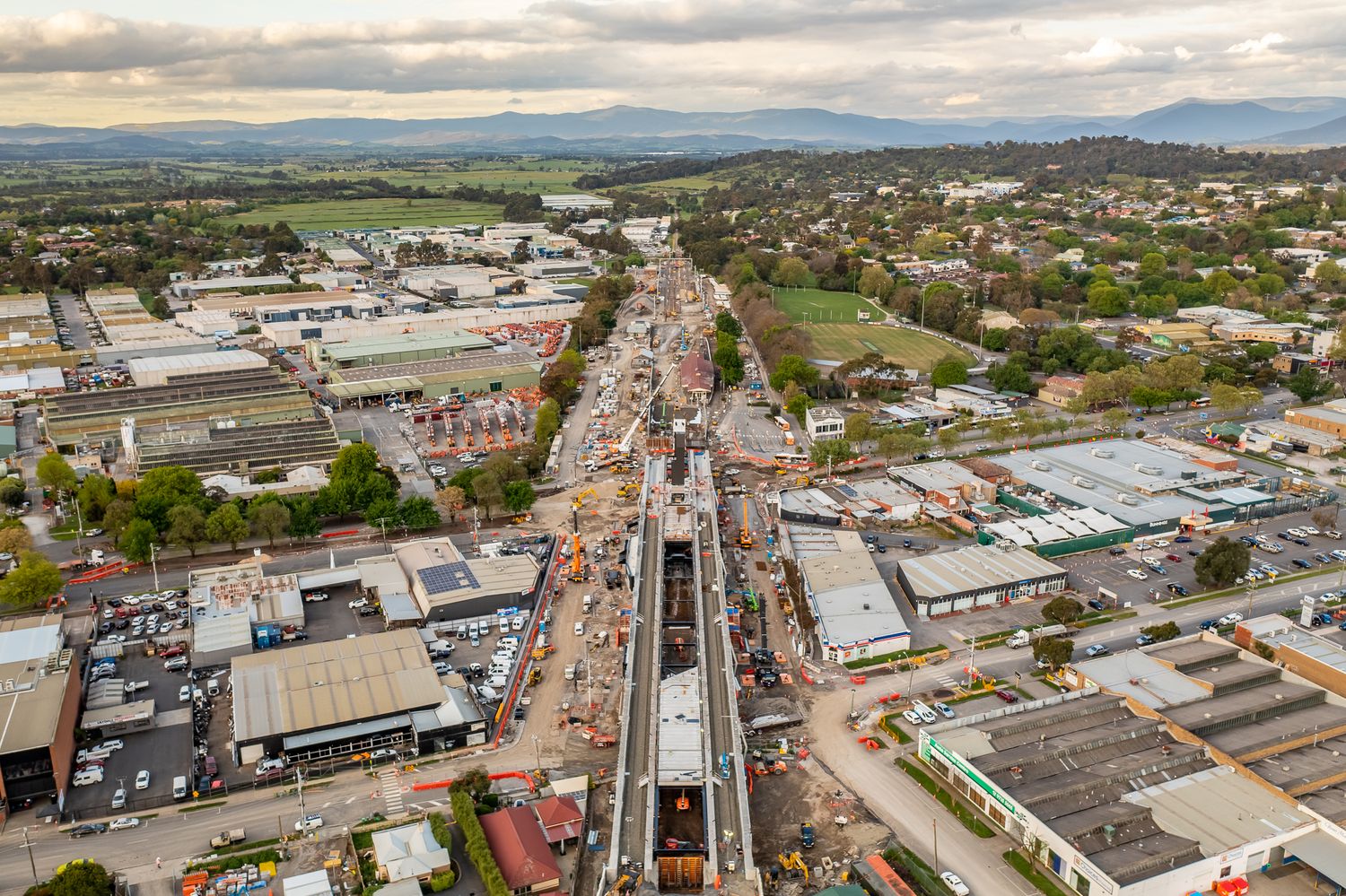 Aerial view of major railway construction project along busy road in Australian suburban town with mountains.