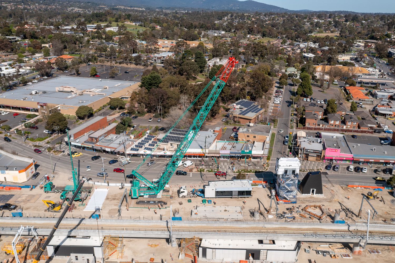 Large green crawler crane lifting materials at busy urban railway construction site in Australian suburb.
