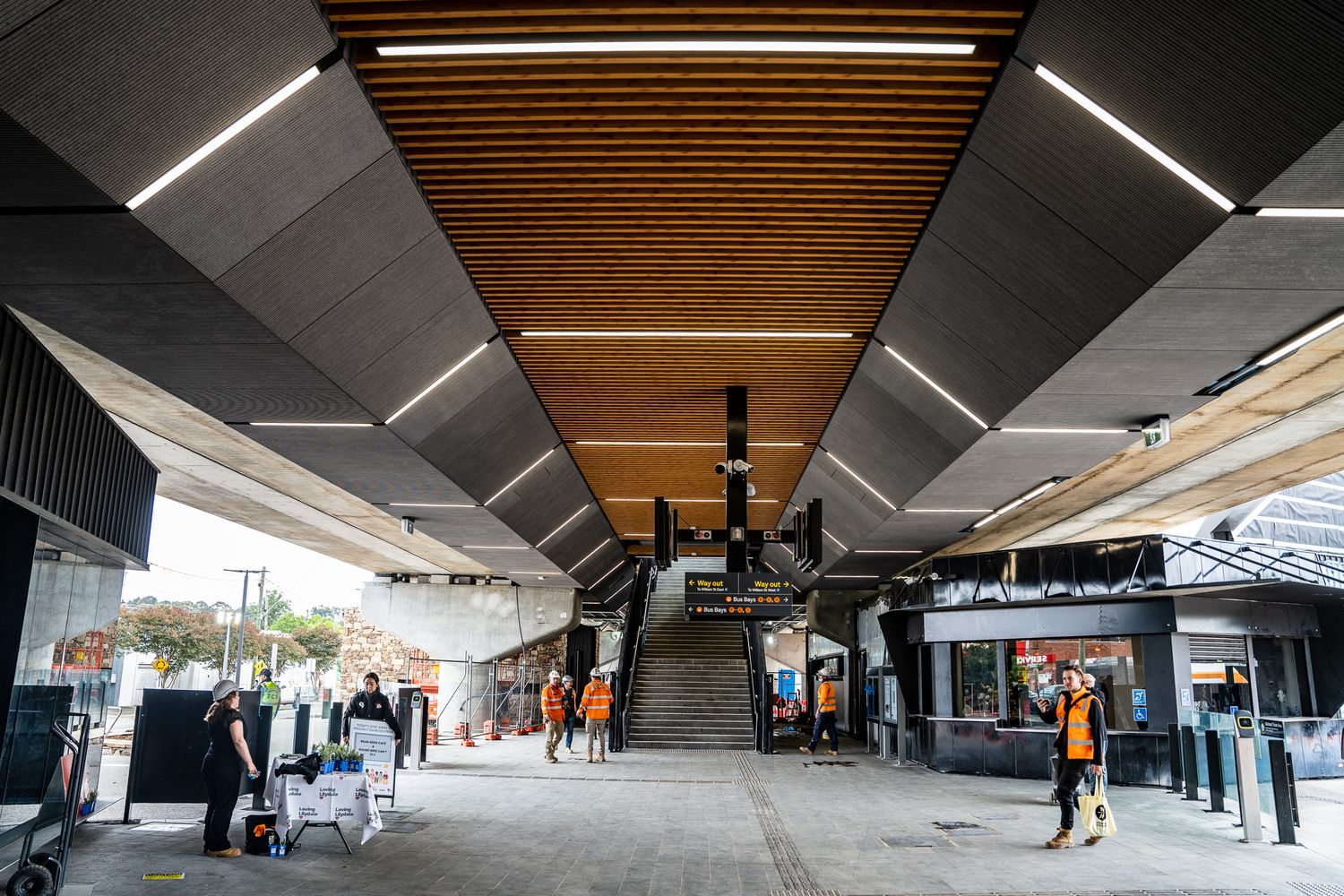 Modern train station concourse with geometric ceiling design, LED lighting strips, and commuters walking below.