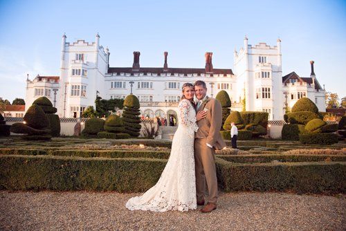Elegant couple poses in front of historic white mansion with manicured gardens.