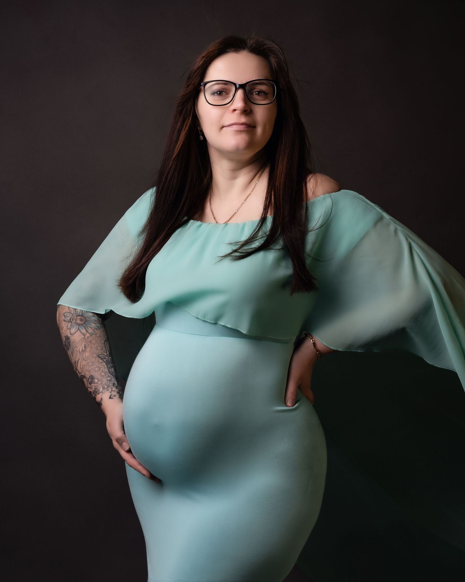 Maternity portrait in flowing mint green dress with butterfly sleeves against dark backdrop.