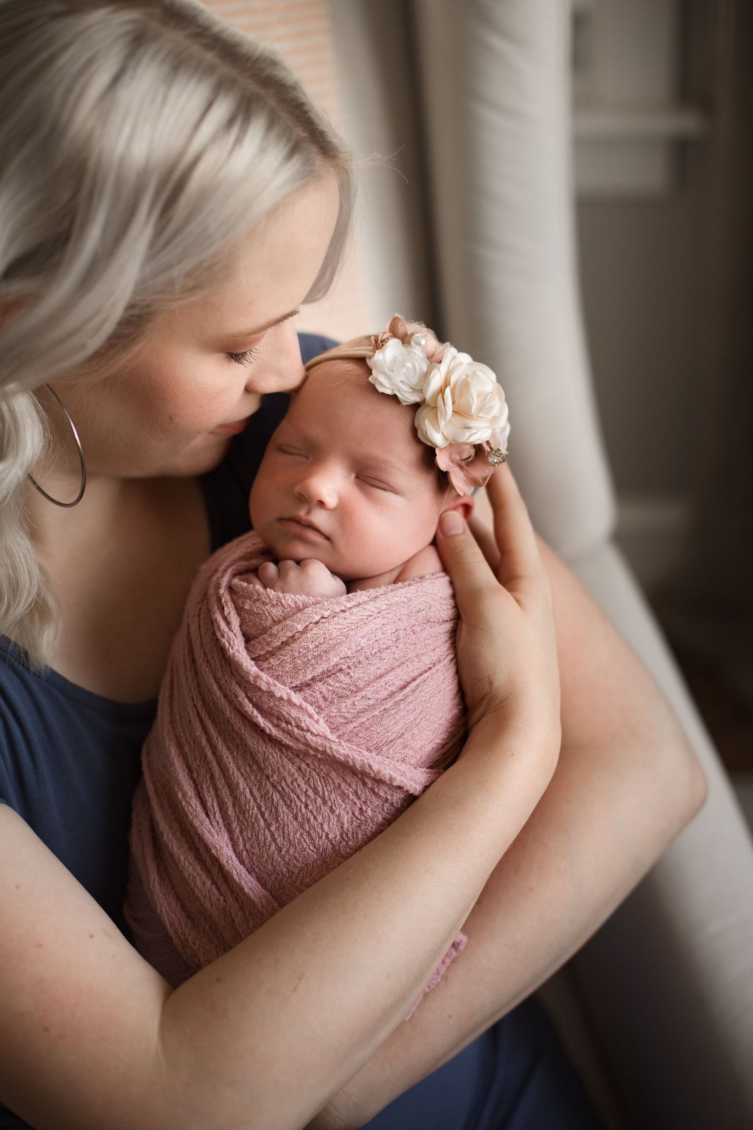 A mother tenderly kisses her swaddled newborn baby girl wearing a floral headband.