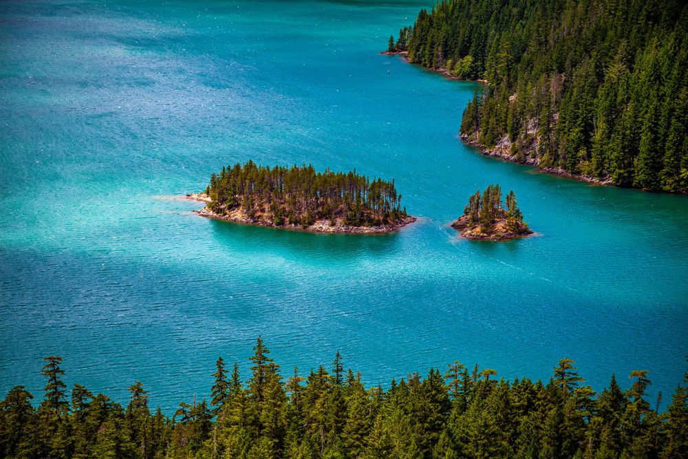Small forested islands dot the turquoise waters of an alpine lake surrounded by evergreen forest.