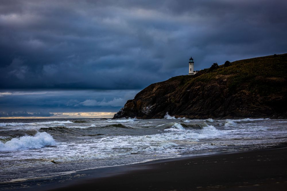 Lighthouse perched on stormy coastal cliff with dramatic waves crashing against dark rocky shore.