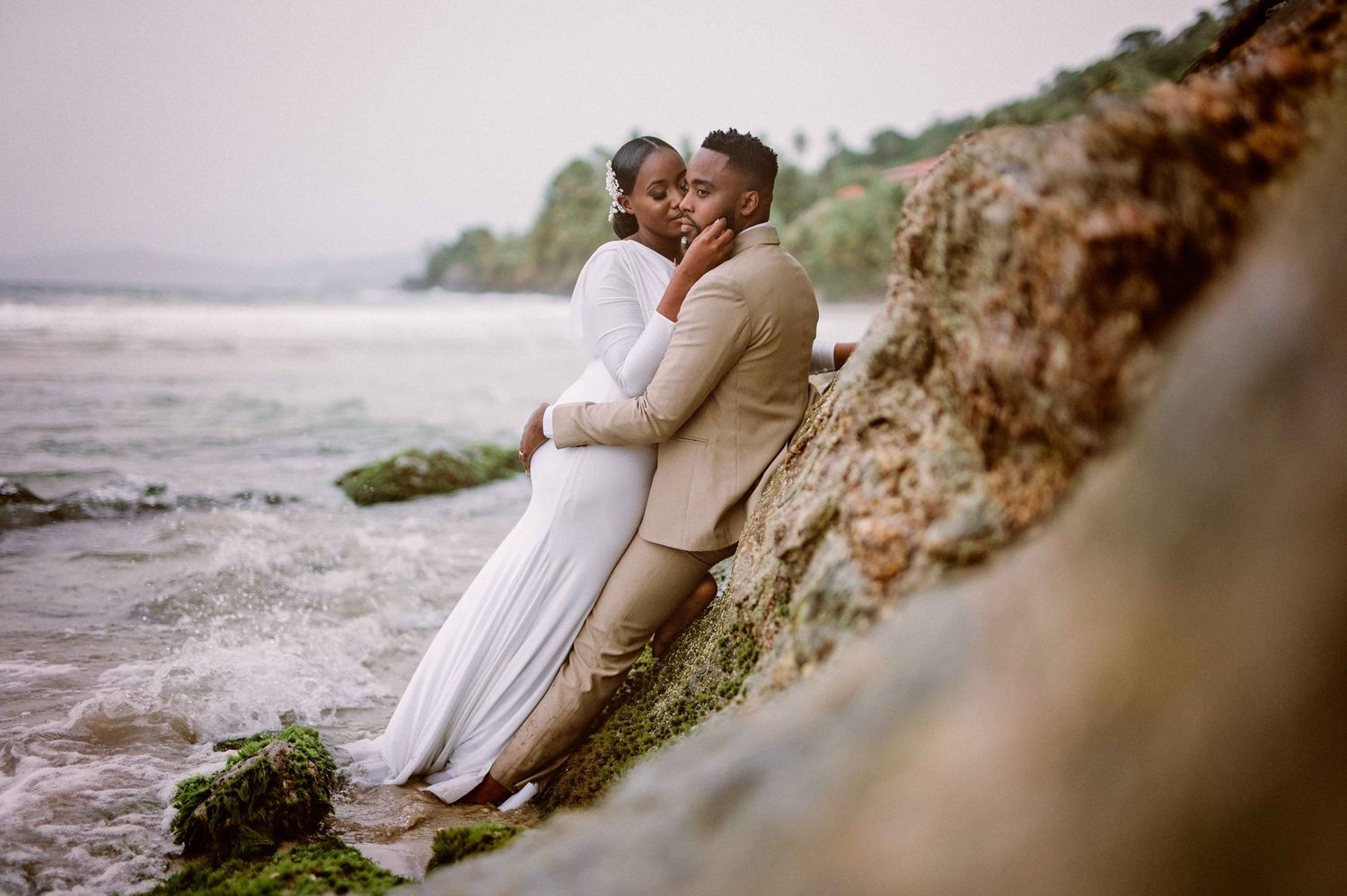 A romantic seaside wedding photo with a couple embracing near rocky cliffs at sunset.