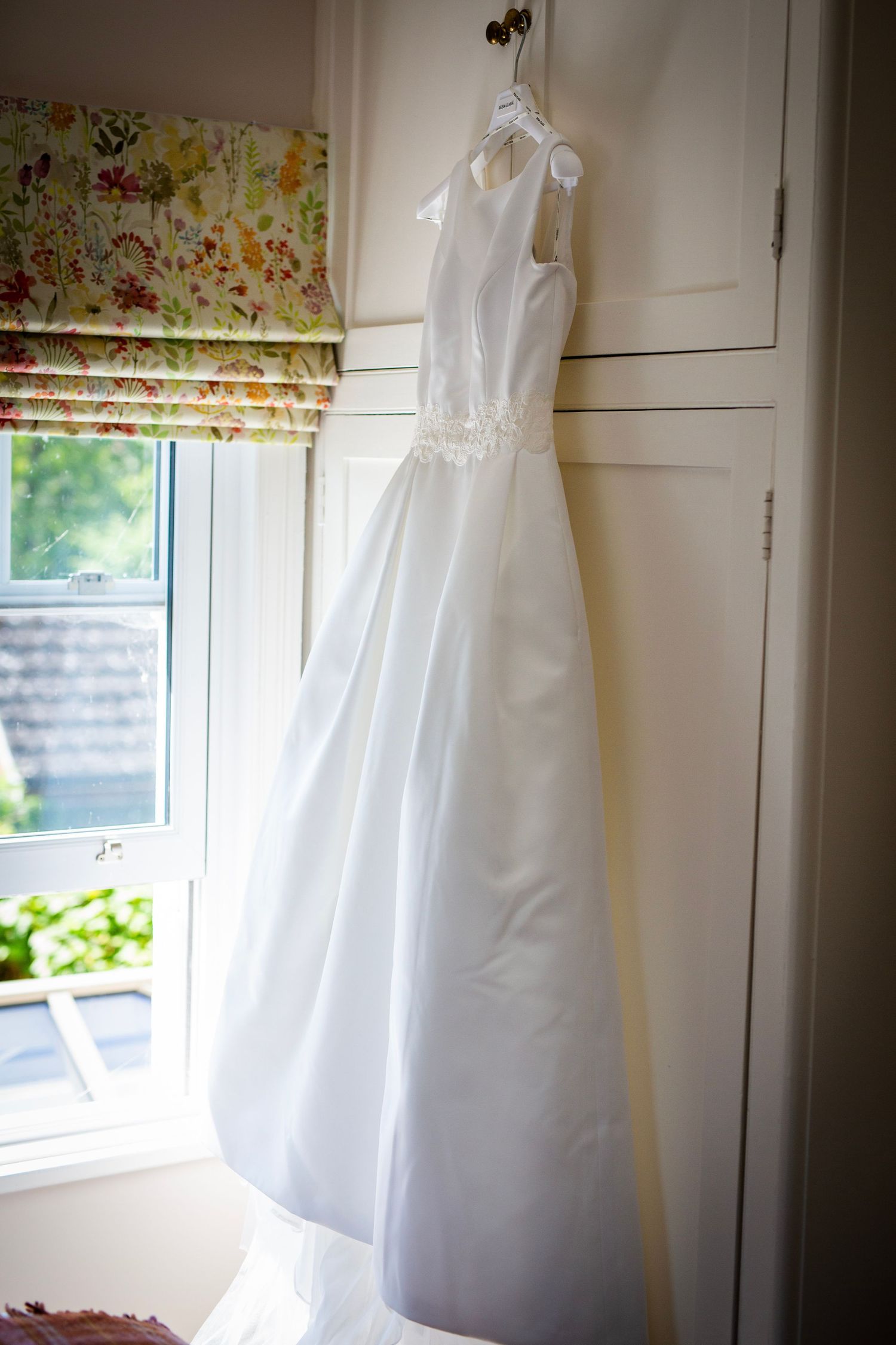 White wedding dress hanging by window with floral curtains in natural light.