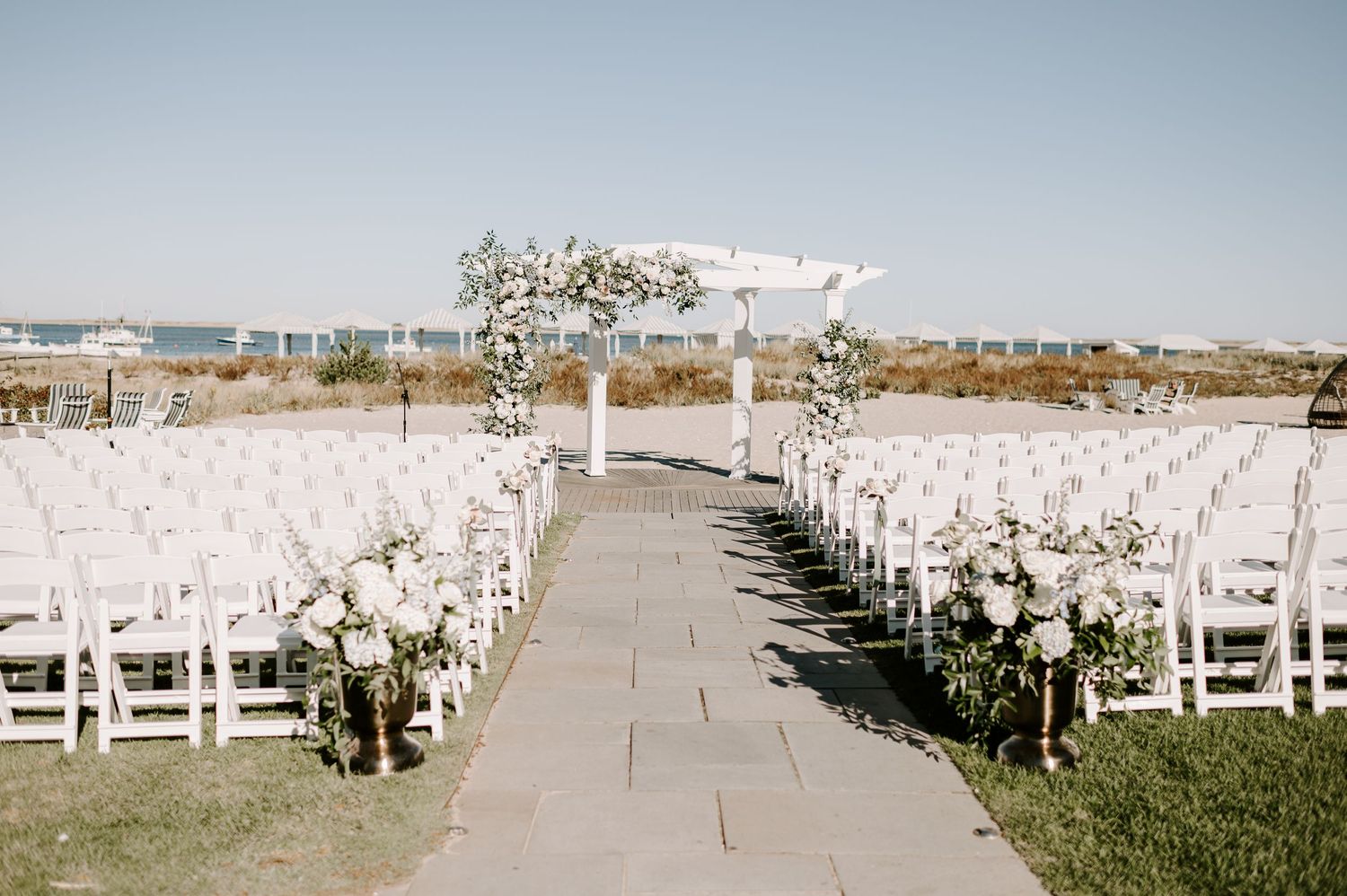 White wedding ceremony setup with flower-decorated arch and chairs lined along stone pathway.