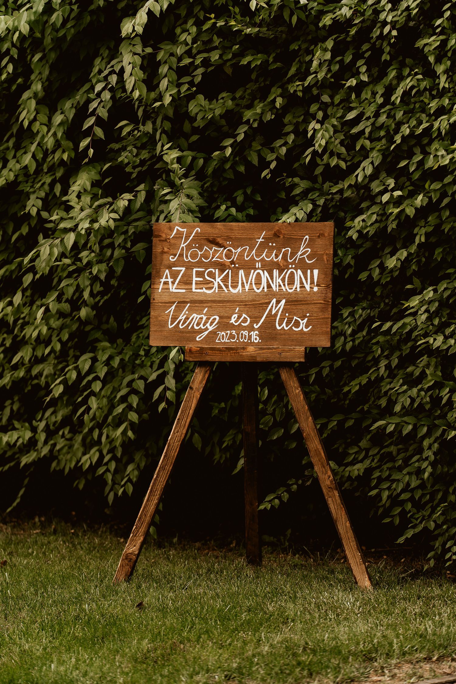 Rustic wooden wedding welcome sign displayed on easel against green leafy backdrop.