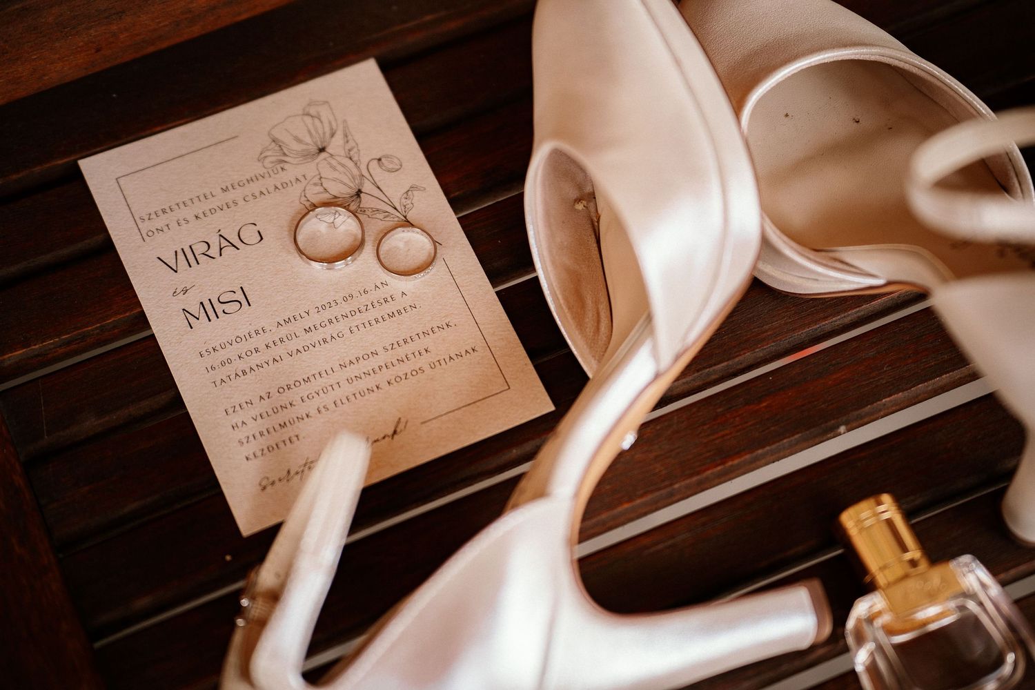 Wedding invitation, white heels, and perfume bottle arranged on dark wood surface.