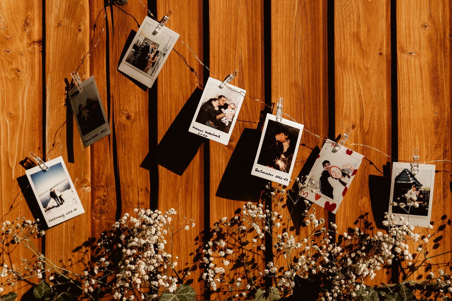 Polaroid photos hanging on string against wooden wall with dried flowers below.