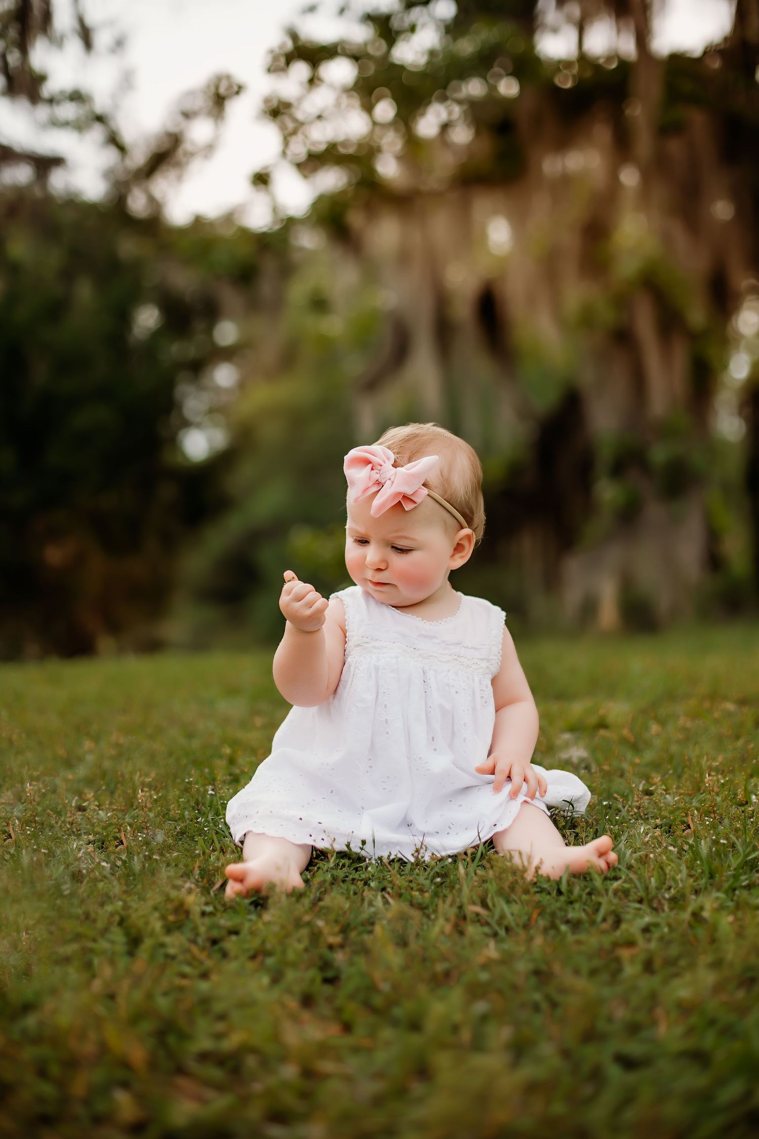 Baby wearing white dress and pink bow sitting on grass outdoors.