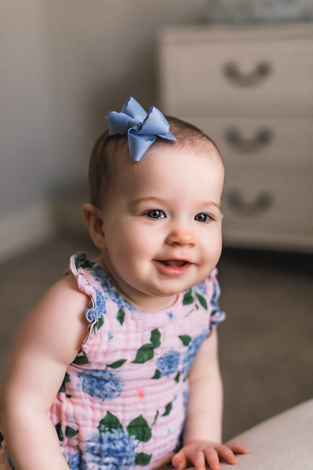 Baby in floral dress and blue bow smiling while leaning on furniture.