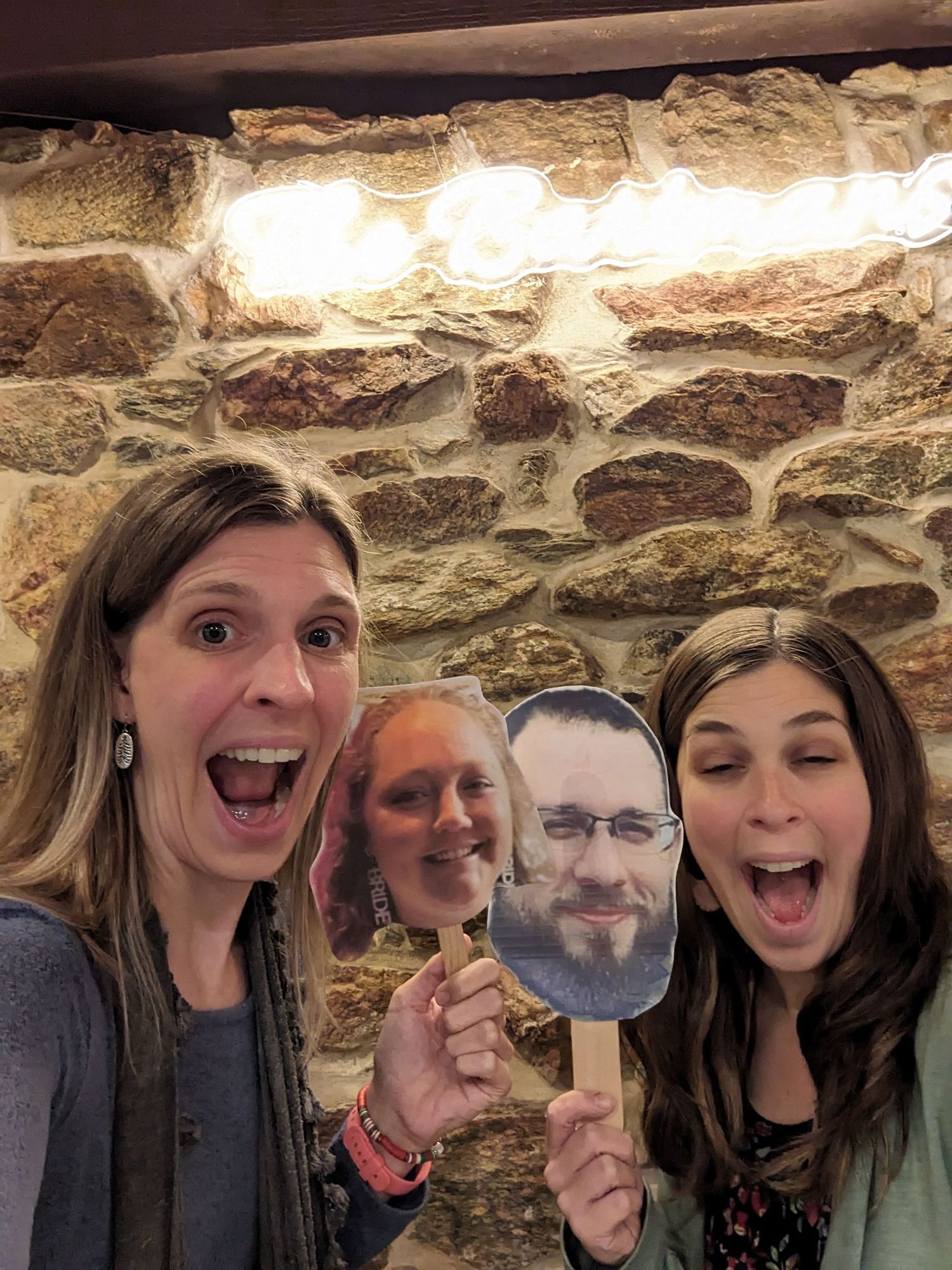Friends making silly expressions while holding a face cutout against a rustic stone wall background.