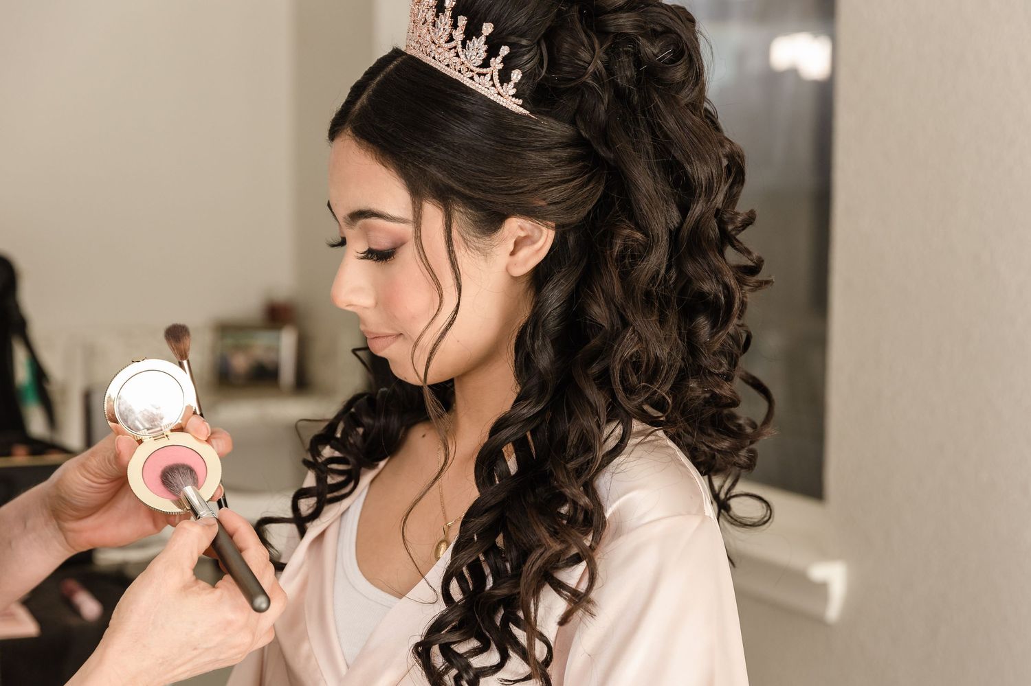 Professional makeup artist applies cosmetics to a woman wearing a tiara and white robe with curled dark hair.