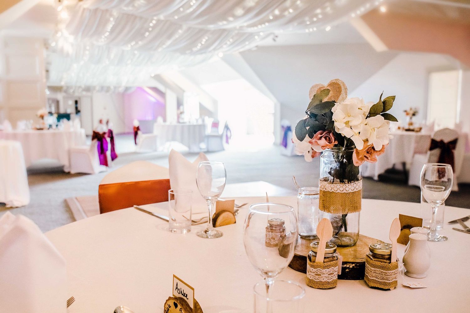 Elegant white wedding reception table setting with floral centerpiece in bright venue.