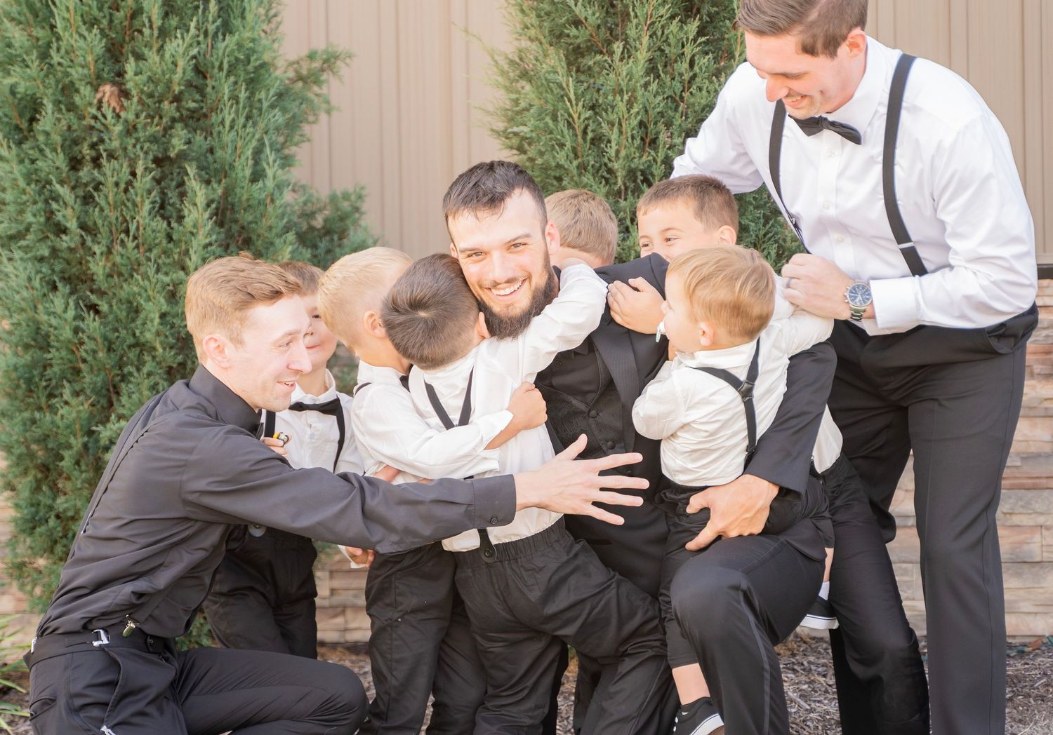 Groom laughing as groomsmen and young ring bearers playfully pile on him outdoors.
