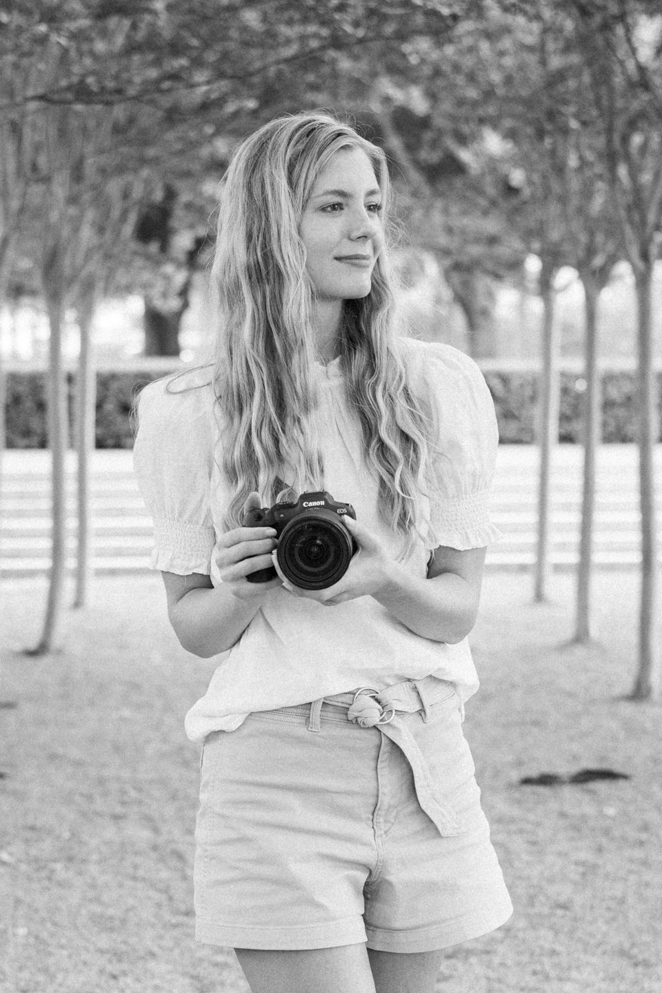 A photographer in casual summer attire holds a camera in a tree-lined setting.
