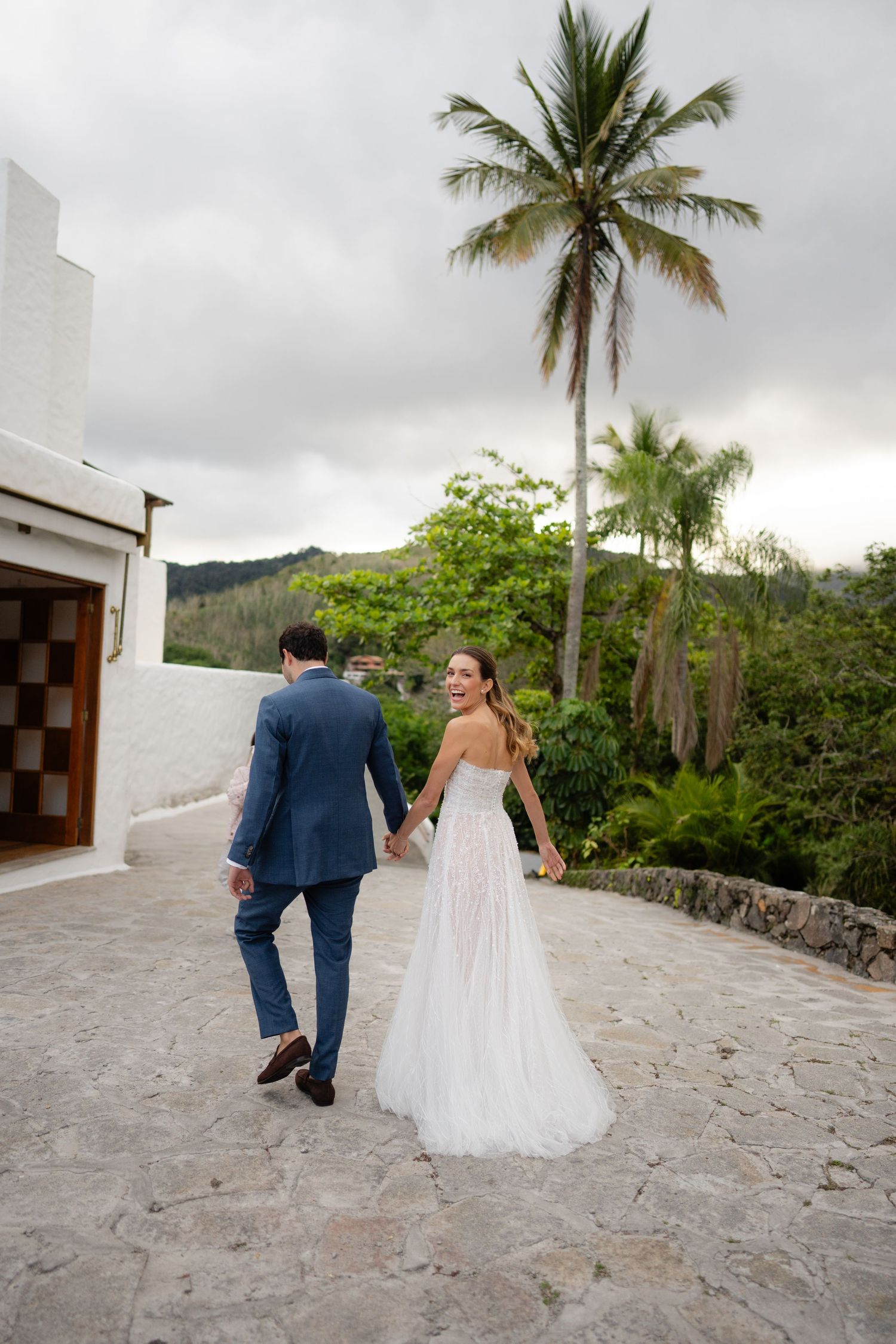 Newlyweds walking hand in hand on stone path with tropical palm trees overhead.