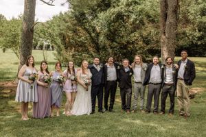 Large wedding party poses together outdoors with bridesmaids in purple dresses and groomsmen in suits.