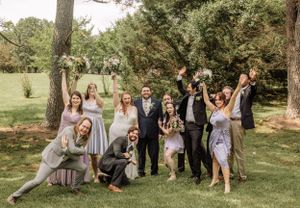 Wedding party members pose informally on grass under mature trees.