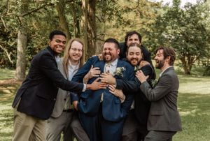 A group of groomsmen laugh and joke together while posing for photos in a wooded outdoor setting.