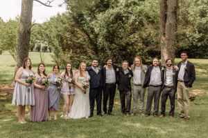 Wedding party poses together outdoors under tall trees in a natural setting with bridesmaids in purple dresses.